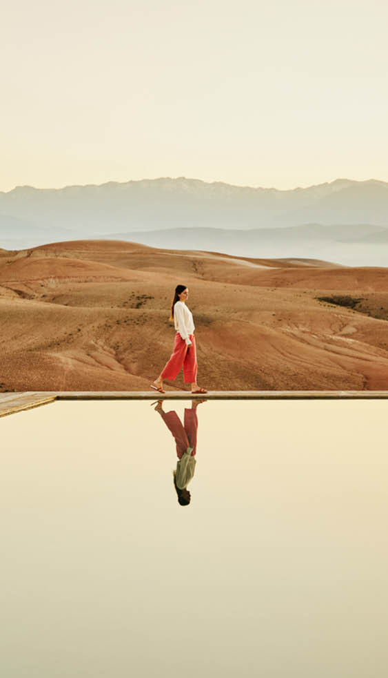 Extreme wide shot of solo female traveler walking along the edge of luxury desert camp swimming pool at sunrise enjoying view of Agafay Desert during vacation in Morocco